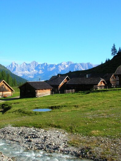 Eine malerische Landschaft mit traditionellen Holzgebäuden und einem kleinen Bach. Im Hintergrund erstrecken sich beeindruckende Berge unter einem klaren blauen Himmel. | © Ursprungalm