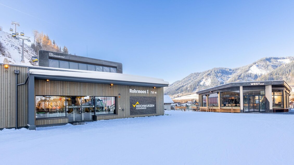 Ein moderner Gebäudekomplex in einer schneebedeckten Berglandschaft. Im Hintergrund sind verschneite Berge unter einem klaren blauen Himmel sichtbar.