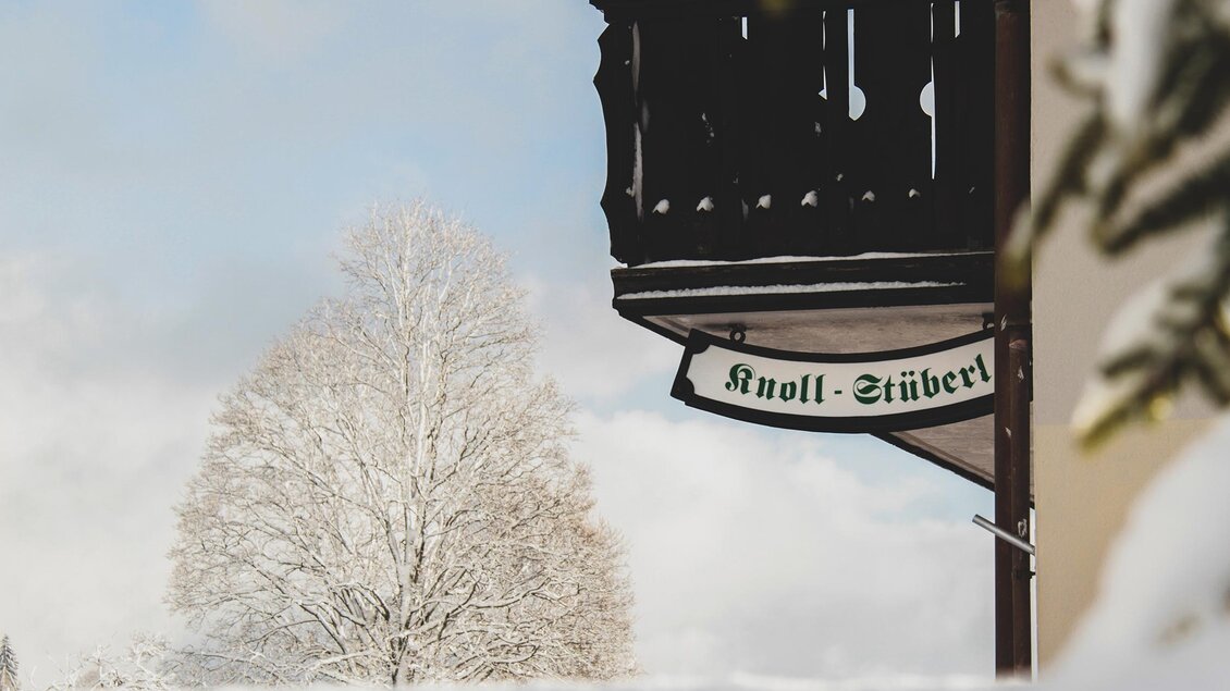 Ein Holzbalkon mit einem Schild sticht bei einer verschneiten Landschaft heraus. Im Hintergrund ist ein mit Schnee bedeckter Baum zu sehen. | © Foto Tom
