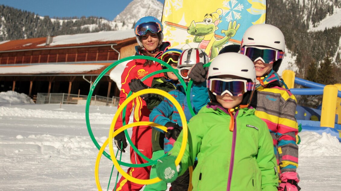 Eine Gruppe von Kindern in bunten Skibekleidungen steht im Schnee. Im Hintergrund sind ein Ski-Lift und ein buntes Plakat zu sehen. | © WM-Schischule Royer