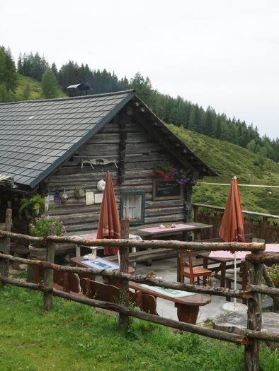 A rustic mountain cabin surrounded by beautiful scenery. Tables with sunshades invite you to relax outdoors. | © Naturpark Sölktäler