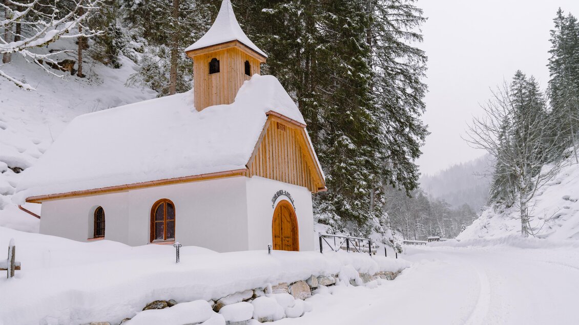 Eine kleine Kapelle im Schnee, umgeben von verschneiten Bäumen. Der Himmel ist grau und winterlich. | © Christoph Lukas