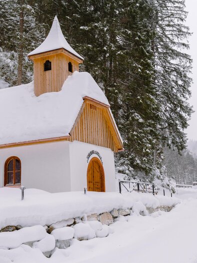 A small chapel in the snow, surrounded by snow-covered trees. The sky is gray and wintry. | © Christoph Lukas
