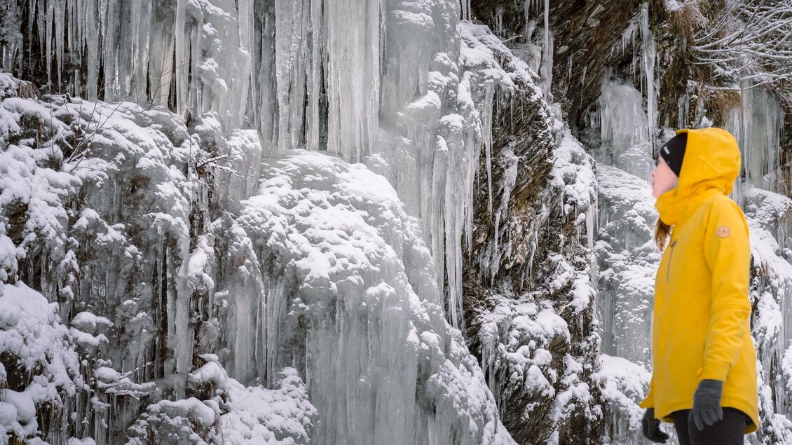 Eine Person in einer gelben Jacke steht vor einer gefrorenen Felswand. Eisige Tropfen hängen herab und der Schnee bedeckt den Boden. | © Christoph Lukas