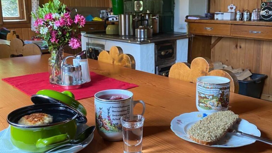 Eine gemütliche Küche mit einem Holztisch, auf dem ein grüner Kochtopf, eine Tasse und ein Stück Brot liegen. Im Hintergrund stehen eine alte Kochzeile und frische Blumen in einer Vase. | © Walhornalm