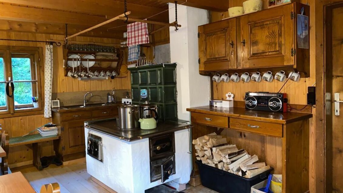 Eine gemütliche Küche im Holzstil mit einem traditionellen Ofen und viel Stauraum. Der Raum ist hell und einladend, mit einem Blick auf die Natur durch das Fenster. | © Walhornalm