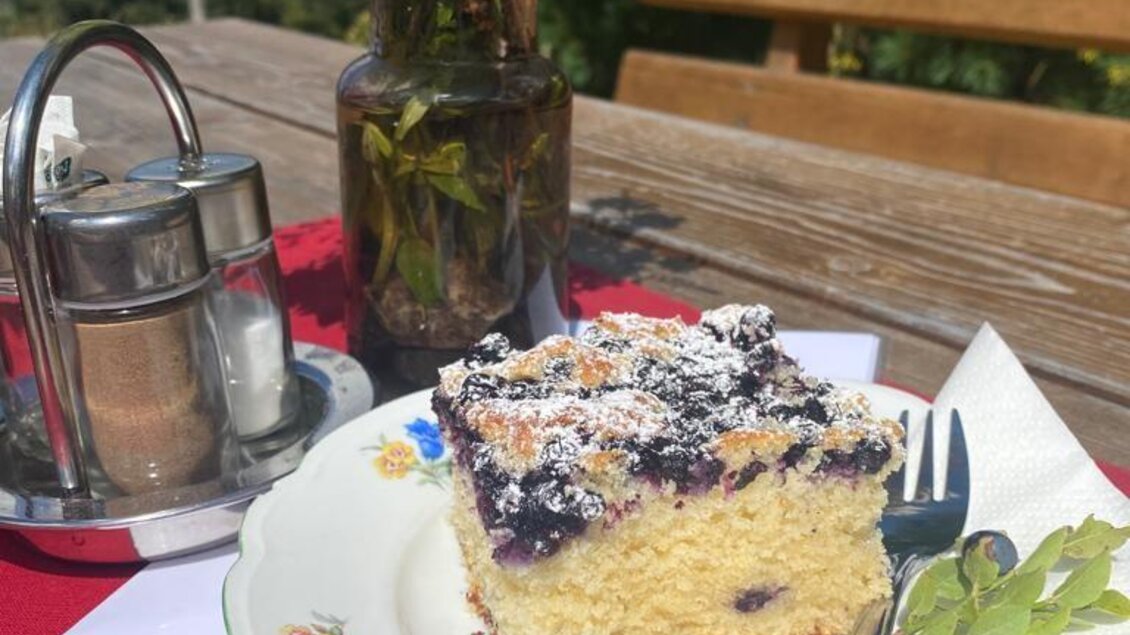 Ein Stück Kuchen mit Blaubeeren und Puderzucker liegt auf einem Teller. Im Hintergrund steht eine Blumenvase auf einem Holztisch. | © Walhornalm