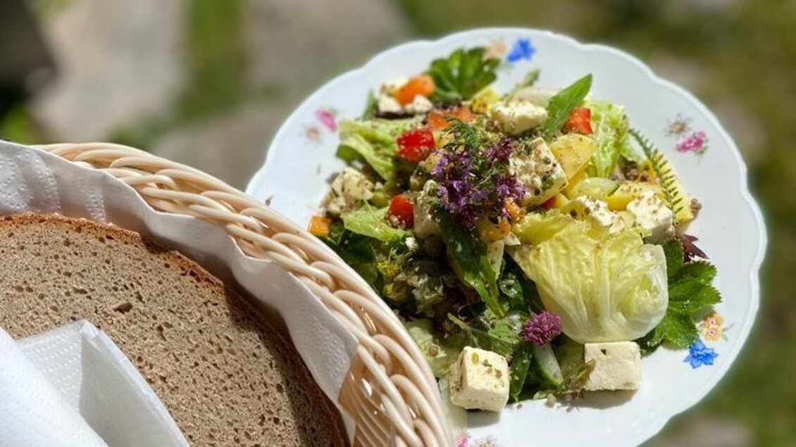Ein bunter Salat mit frischem Gemüse und Feta-Käse auf einem weißen Teller. Daneben ein Korb mit dunklem Brot und Serviette. | © Walhornalm