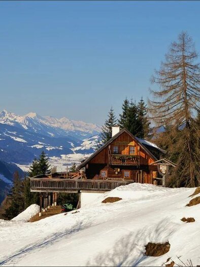 Eine gemütliche Berghütte steht auf einem schneebedeckten Hang. Im Hintergrund sind majestätische Berge und ein klarer blauer Himmel zu sehen. | © Wiesmahdalm