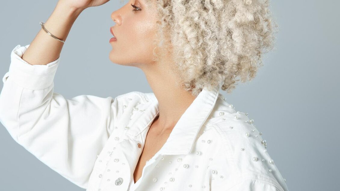 Eine Frau mit lockigem, blondem Haar trägt eine weiße Jacke. Sie schaut nachdenklich zur Seite. | © Wohlfühlfriseur Rohrmoser