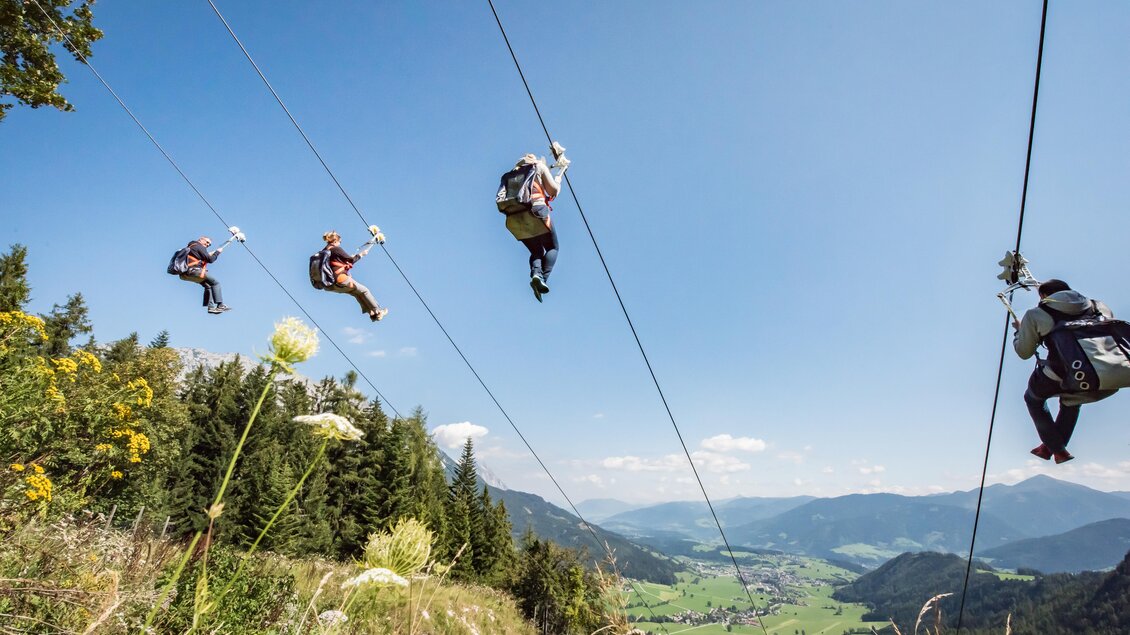 Fünf Personen fahren an einer Zipline über eine malerische Landschaft. Im Hintergrund sind Berge und ein klarer blauer Himmel zu sehen. | © Christoph Huber