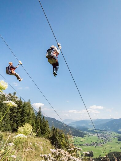 Five people are riding on a zipline over a picturesque landscape. In the background, there are mountains and a clear blue sky. | © Christoph Huber