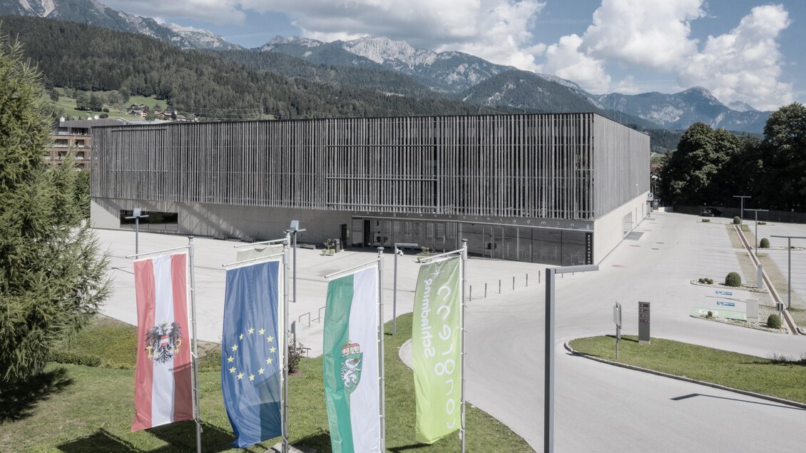 Ein modernes Gebäude umgeben von grüner Landschaft und Bergen. Vor dem Gebäude wehen mehrere Fahnen im Vordergrund. | © Harald Steiner