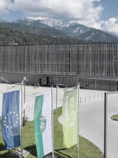 A modern building surrounded by green landscape and mountains. In front of the building, several flags are waving in the foreground. | © Harald Steiner