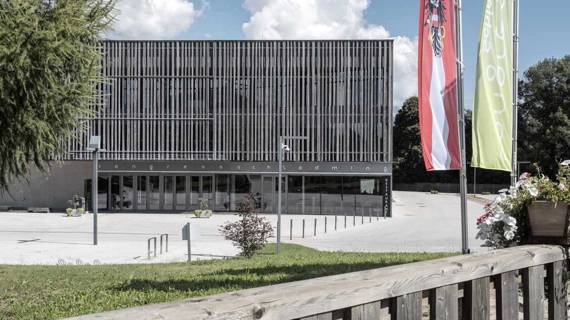 Ein modernes Gebäude mit Holzlattenfassade und großen Glasfronten. Im Vordergrund sind Fahnen und eine grüne Wiese zu sehen. | © Harald Steiner