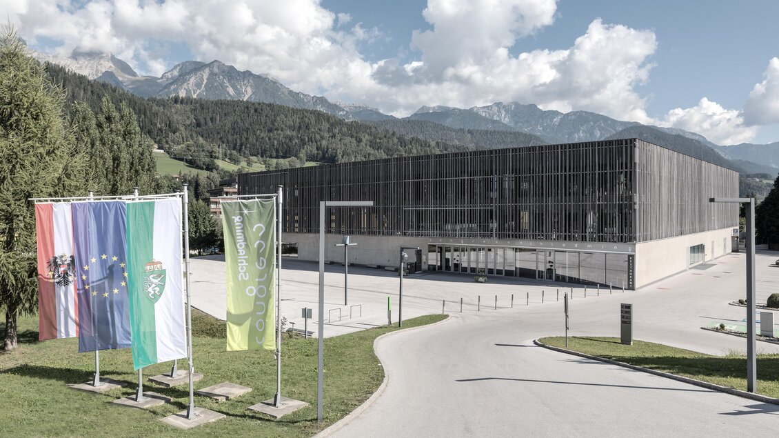 Ein modernes Gebäude mit einer großen Glasfassade und umliegenden Wäldern. Im Vordergrund stehen Flaggen und die beeindruckende Berglandschaft ist im Hintergrund sichtbar. | © Harald Steiner