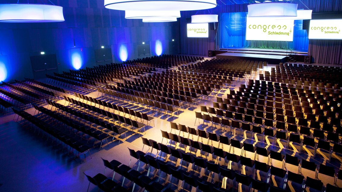 Eine große Konferenzhalle mit reihenweise leeren Stühlen. Die Wände sind beleuchtet und es gibt eine Bühne im Hintergrund. | © Harald Steiner 