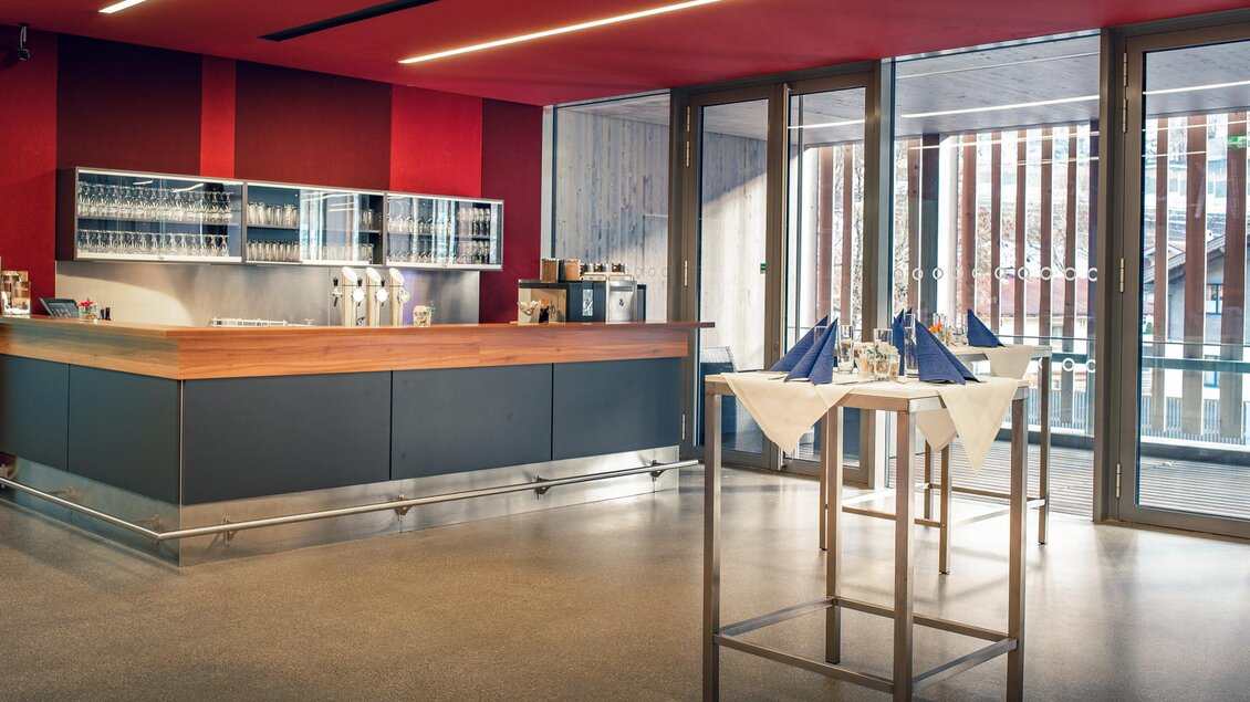 Eine moderne Bar mit einem Holztresen und einer eleganten Einrichtung. Die großen Fenster lassen viel Licht herein und bieten einen Blick nach draußen. | © congress Schladming