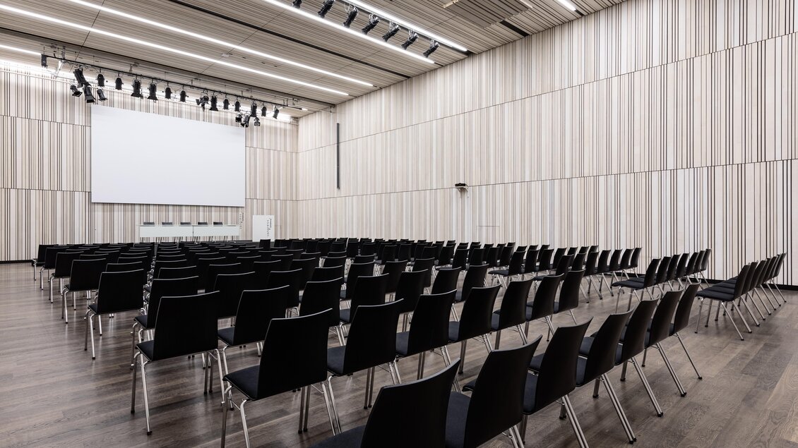 Ein moderner Veranstaltungsraum mit vielen schwarzen Stühlen und einem großen Bildschirm an der Wand. Der Raum hat eine helle, minimalistische Gestaltung. | © Harald Steiner 