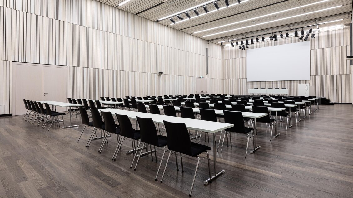 Ein moderner Konferenzraum mit mehreren Reihen von Tischen und Stühlen. Eine große Leinwand ist an der Vorderwand angebracht. | © Harald Steiner 