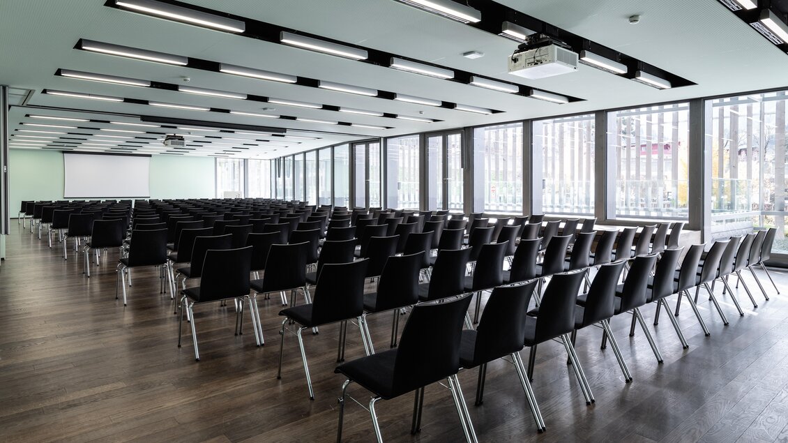 Ein großer, moderner Konferenzraum mit vielen schwarzen Stühlen in Reihen. Große Fenster sorgen für viel natürliches Licht. | © Harald Steiner 
