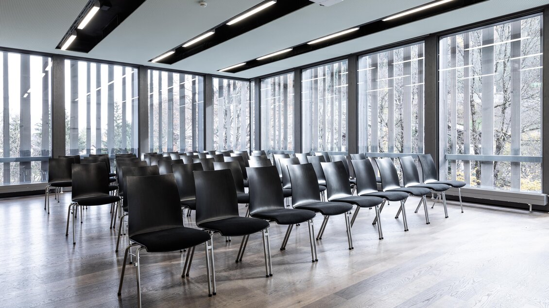 Ein moderner Seminarraum mit vielen schwarzen Stühlen in Reihen. Große Fenster bieten viel Tageslicht und einen Blick nach draußen. | © Harald Steiner 