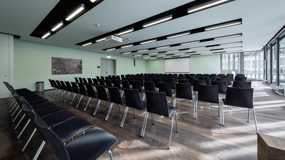 Ein moderner Konferenzraum mit vielen Stühlen in aufgereihter Ordnung. Der Raum hat große Fenster und eine Präsentationsleinwand an der Wand. | © Harald Steiner 