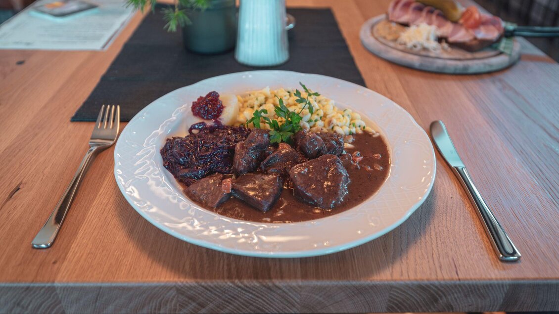 Ein Teller mit zartem Fleisch in einer dunklen Soße, serviert mit Reis und Preiselbeersauce. Im Hintergrund ist eine andere Platte mit Schinken und weiteren Speisen zu sehen. | © d´Genussalm