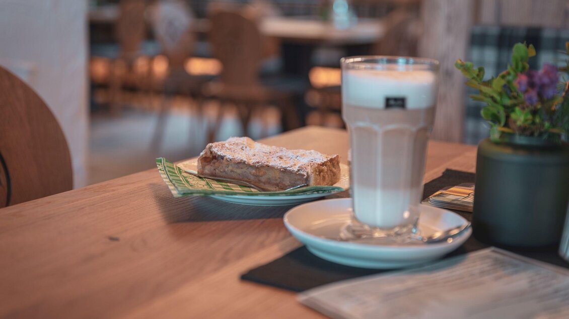 Ein Glas Kaffee steht neben einem Stück Kuchen auf einem Tisch. Die Umgebung wirkt gemütlich und einladend. | © d´Genussalm