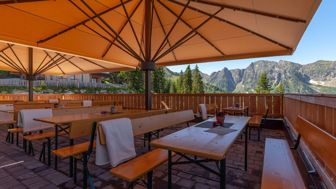 Eine gemütliche Außenbereich mit Holztischen und Bänken unter schattigen Sonnenschirmen. Im Hintergrund sind beeindruckende Berge und ein klarer Himmel sichtbar. | © d´Genussalm