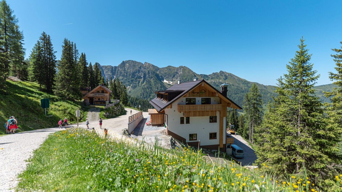 Eine malerische Berglandschaft mit einem Holzhaus und grünen Wiesen. Im Hintergrund sind hohe Berge und ein klarer Himmel zu sehen. | © d´Genussalm