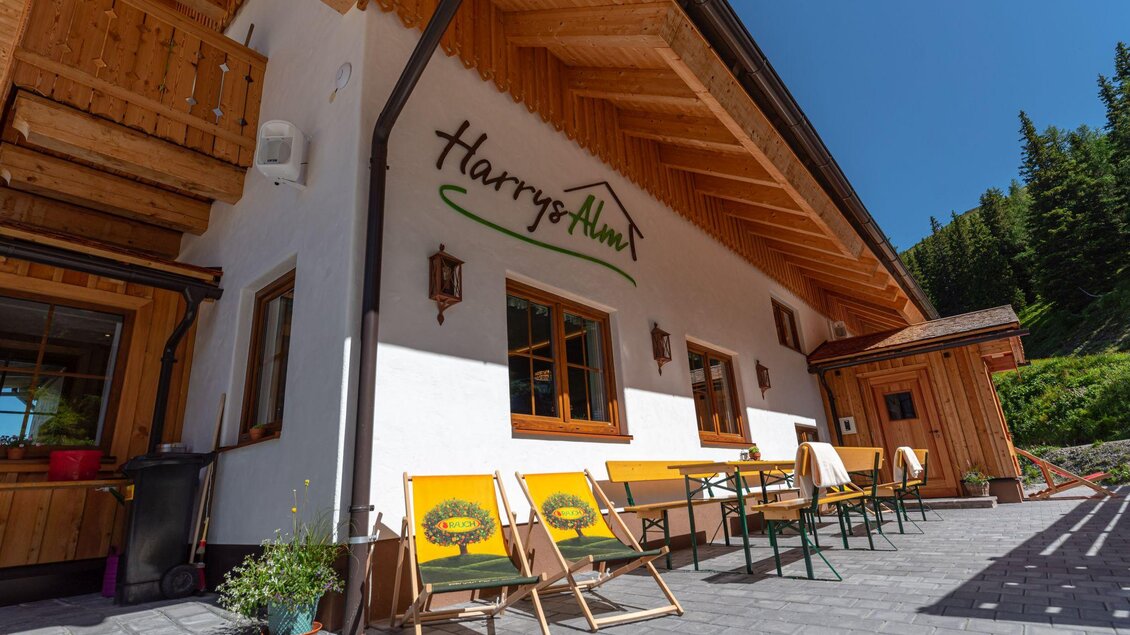 Ein gemütliches Gebäude mit Holzverkleidung und großen Fenstern. Vor dem Eingang stehen Sonnenliegen und Tische in einer schönen Umgebung. | © d´Genussalm