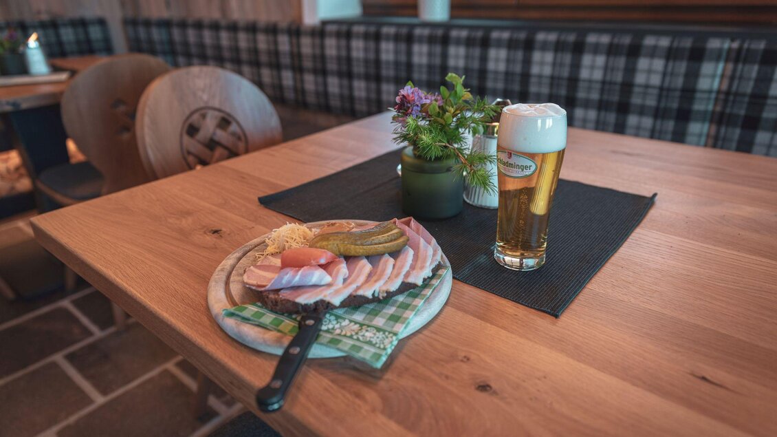 Ein Holztisch mit einem Teller Wurst und Käse sowie einem Glas Bier. Im Hintergrund sind gemütliche Sitzgelegenheiten zu sehen. | © d´Genussalm