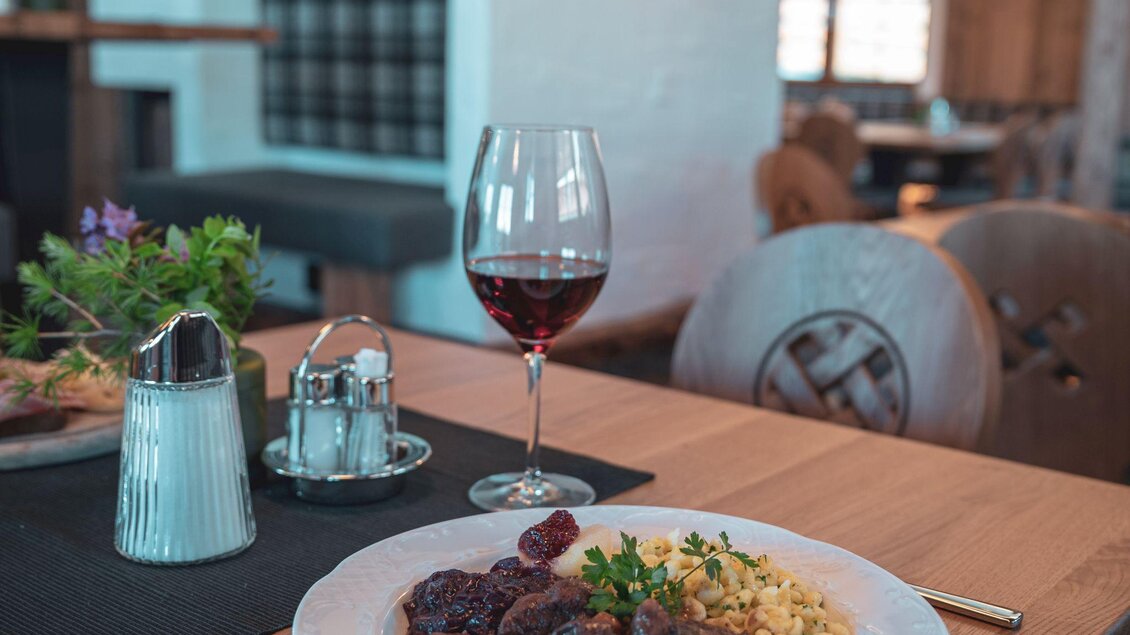 Ein elegantes Restaurant mit einem schön gedeckten Tisch. Auf dem Teller liegt ein Gericht mit Fleisch, Beilagen und einem Glas Rotwein. | © d´Genussalm