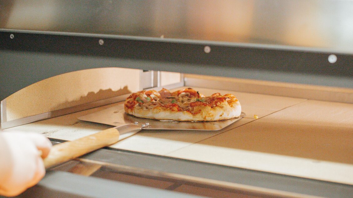 Eine frisch gebackene Pizza wird aus dem Ofen genommen. Der Käse ist geschmolzen und die Zutaten sind gut sichtbar. | © Daniela Pattermann