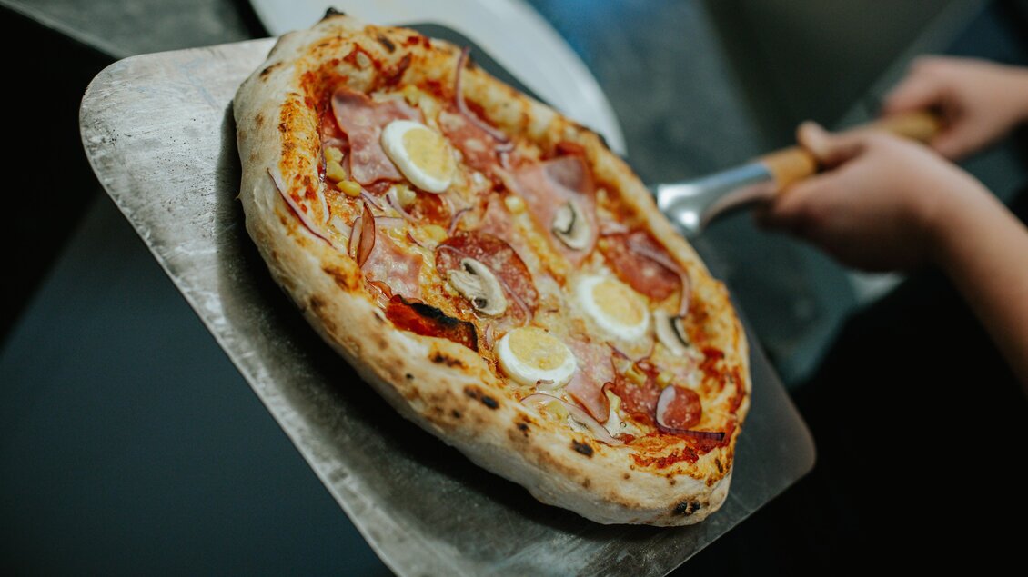 Eine frisch gebackene Pizza mit Schinken, Pilzen und Eiern. Sie wird gerade aus dem Ofen auf einen Pizzaschieber gehoben. | © Daniela Pattermann