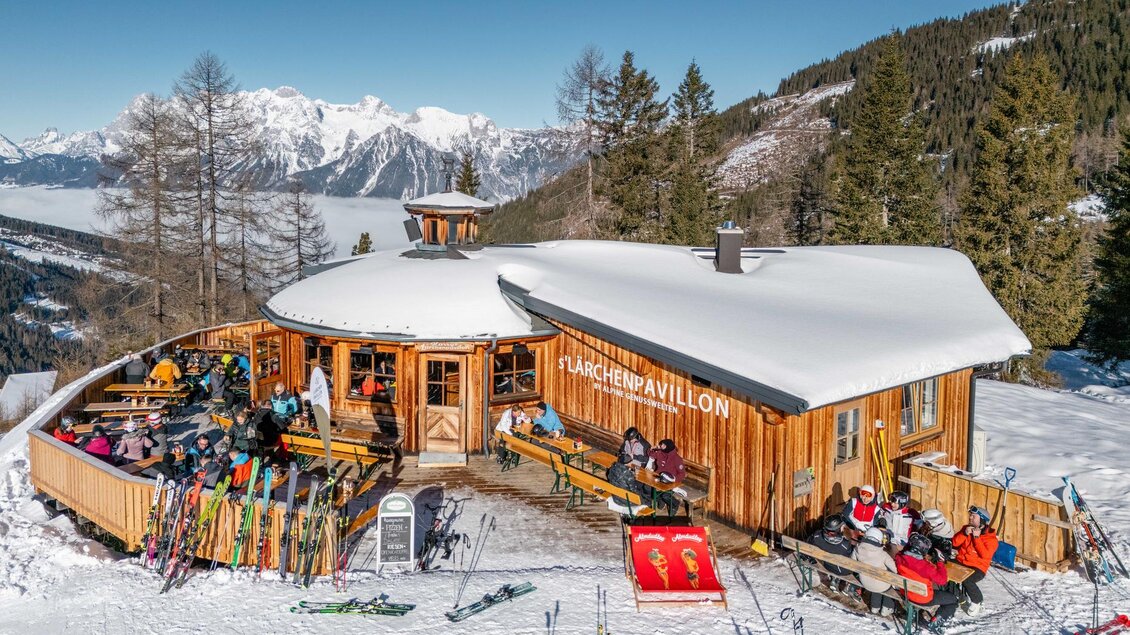 Ein gemütliches Berghaus mit vielen Gästen auf der Terrasse, umgeben von schneebedeckten Bergen. Die Stimmung ist lebhaft und einladend. | © s´Lärchenpavillon