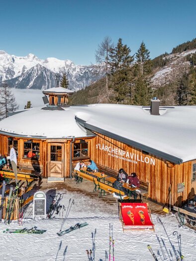 Ein gemütliches Berghaus mit vielen Gästen auf der Terrasse, umgeben von schneebedeckten Bergen. Die Stimmung ist lebhaft und einladend. | © s´Lärchenpavillon