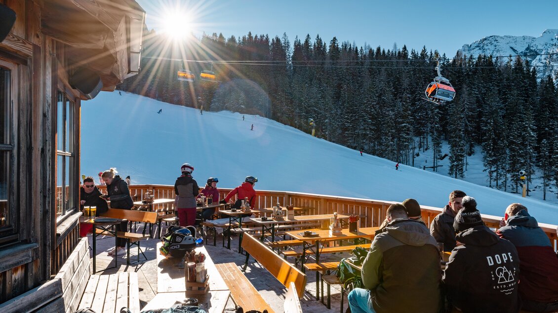 Eine sonnige Skihütte mit Gästen auf einer Terrasse. Im Hintergrund ist eine schneebedeckte Piste und eine Gondel zu sehen. | © s´Lärchenpavillon