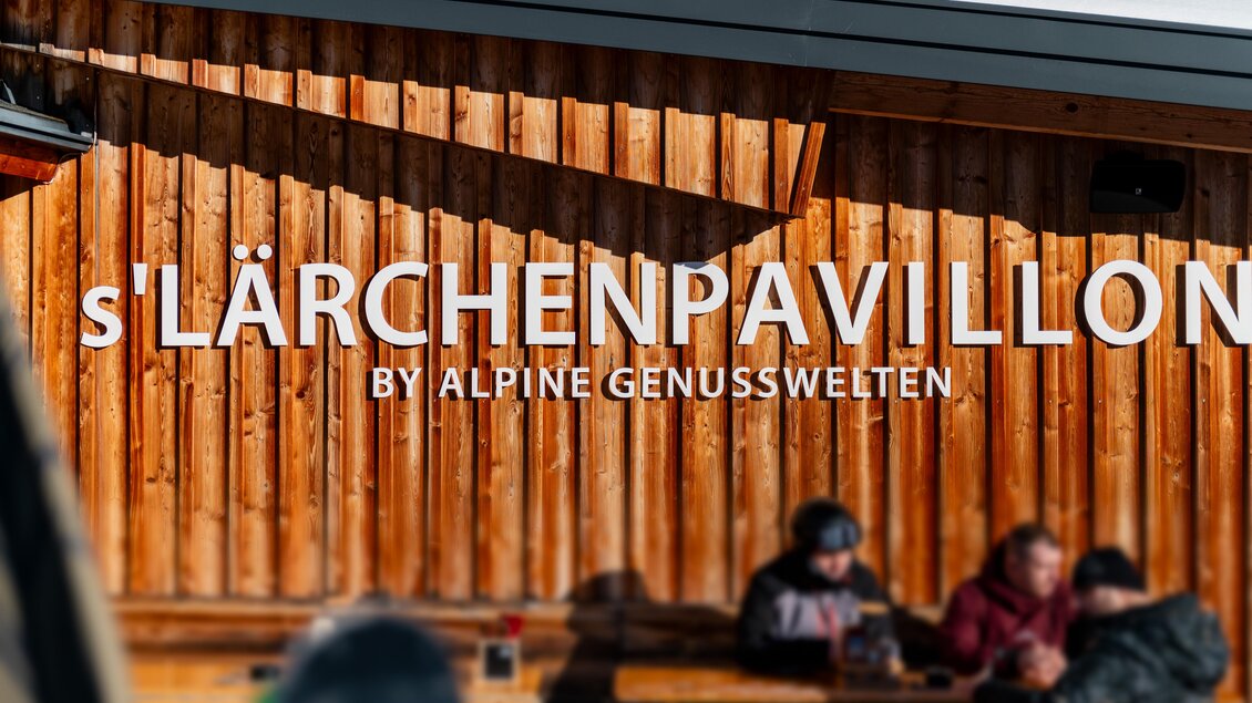 Eine Holzfassade mit der Aufschrift „s'Lärchenpavillon by Alpine Genusswelten“. Im Vordergrund sind unscharfe Personen, die an einem Tisch sitzen und sich unterhalten. | © s´Lärchenpavillon