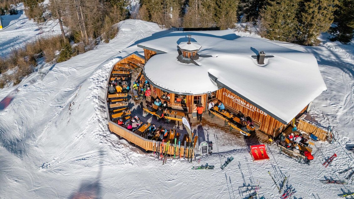 Eine gemütliche Berghütte im Schnee mit vielen Gästen. Umgeben von Bäumen und Skiausrüstung auf der Terrasse. | © s´Lärchenpavillon