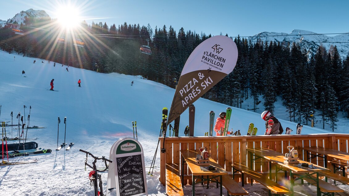 Ein sonniger Skihang mit zahlreichen Skifahrern und einer schönen Aussicht auf die Berge. Im Vordergrund befindet sich eine Terrasse mit Tischen und einem Schild für Pizza und Après Ski. | © s´Lärchenpavillon