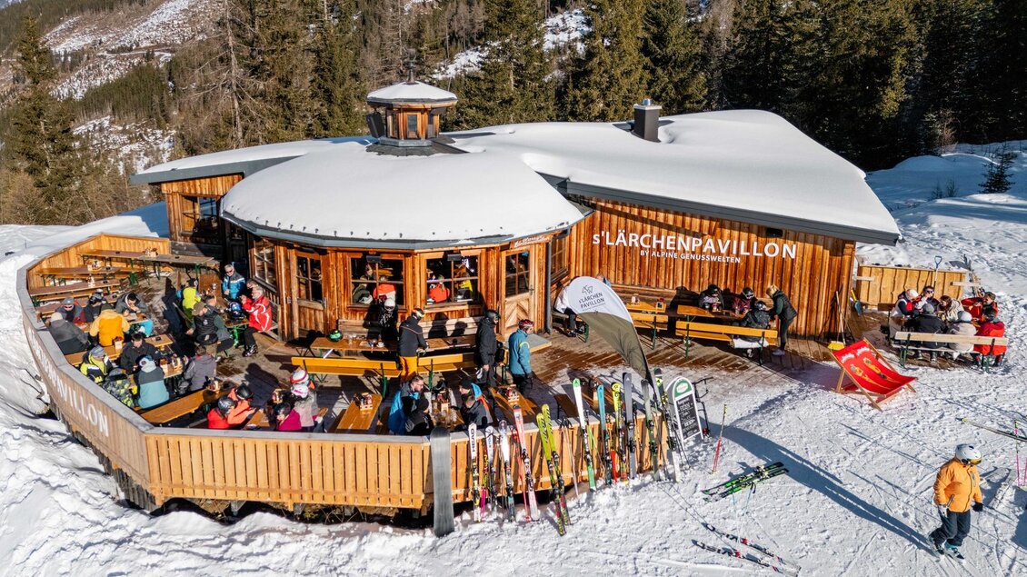 Ein beliebtes Berghaus mit einer großen Terrasse, auf der Skifahrer entspannen. Die Umgebung ist schneebedeckt und von Bäumen umgeben. | © s´Lärchenpavillon