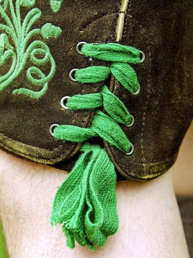 Ein Teil einer traditionellen Tracht mit grünen Schnürsenkeln und einem verzierten Stoff. Die Sicht zeigt den oberen Teil eines Beins. | © s´Gwandl